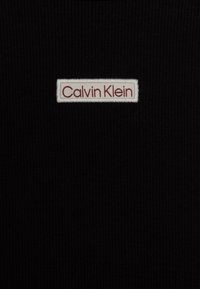 Tessuto nero a coste con un'etichetta bianca centrale che riporta la scritta "Calvin Klein" in lettere bordeaux. La texture è liscia e aderente.