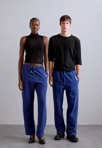 Two models stand side by side, wearing black tops and blue sweatpants. The pants feature an elastic waistband and drawstring.