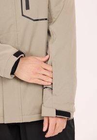 Beige jacka med en strömlinjeformad design, som har en ficka på sidan med dragkedja och kardborreaccentuerade ärmslut. Slät, texturerad tyg.