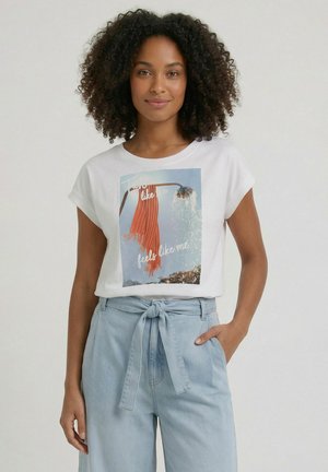 Vrouw met krullend haar die een wit T-shirt draagt met een afbeelding van een douche en sjaal en lichtblauwe high-waisted jeans met een gestrikte riem.