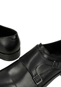 Zwarte leren instapschoenen met dubbele zilveren gespen en lage hakken, getoond vanuit voor- en achterkant op een witte achtergrond.