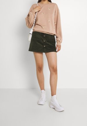 Sweatshirt rose clair avec broderie florale sur le devant, associé à une mini-jupe en velours côtelé vert foncé avec des boutons sur le devant. Baskets montantes blanches.