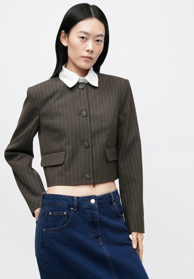 Fiatal nő hosszú fekete hajjal, barna csíkos rövidített zakót visel fehér gallérral és sötétkék, magas derekú farmer szoknyát.