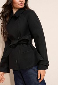 Musta vyöllinen takki, joka on valmistettu teksturoidusta kankaasta. Siinä on kaulus, nappikoristeet ja etutasku. Päällä on tummansiniset farkut.