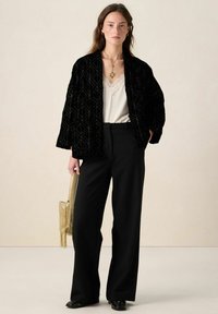 Zwarte fluwelen jas met geometrisch patroon, gedragen over een crème topje met kanten rand, gecombineerd met op maat gemaakte zwarte broek en gouden handtas.
