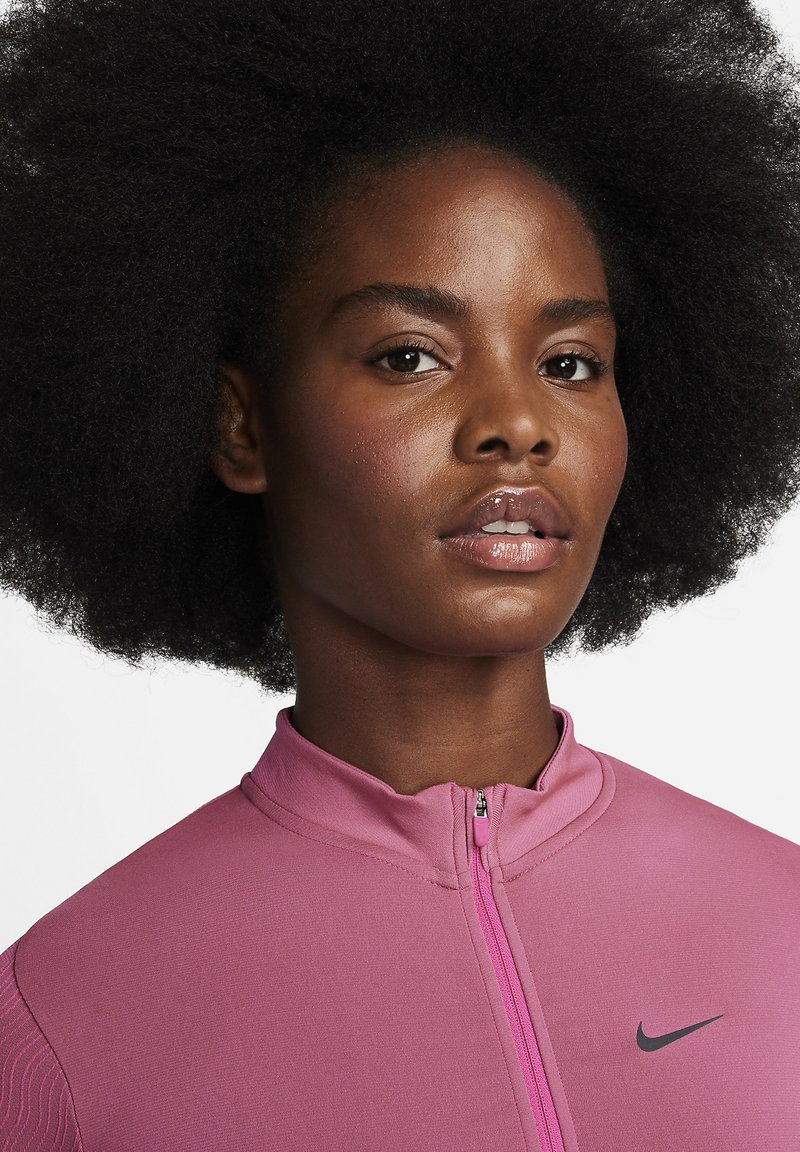 Primer plano de una mujer con cabello afro natural que lleva una chaqueta deportiva rosa con cremallera de Nike, mirando ligeramente hacia un lado con una expresión neutral.