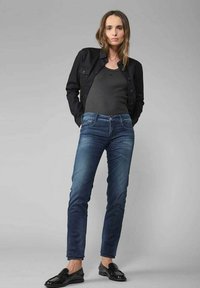 Veste en denim noir sur un débardeur gris, associée à un jean skinny bleu. La tenue comprend des chaussures noires brillantes et présente des détails subtils au niveau des poches.