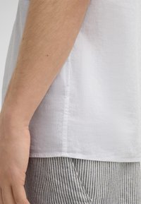 Weißes Leinenhemd mit strukturiertem Finish, gerade geschnitten und mit Nähten an der Seite, kombiniert mit blau-weiß gestreiften Hosen.