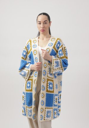 Kobieta o ciemnych włosach nosi długi, oversize'owy kardigan szydełkowy z niebieskimi, białymi i musztardowymi geometrycznymi wzorami, nałożony na biały top i beżowe spodnie.