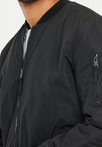 Giacca bomber nera con finitura liscia, collo a costine, zip frontale e tasca con cerniera sulla manica sinistra. Design semplice e funzionale.