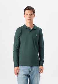 Vihreä pitkähihainen polo paita, jossa on kaulus, ja jossa on pieni logo rinnassa, yhdistettynä vaaleansinisiin farkkuihin. Pehmeä, tekstuuriinen kangas.