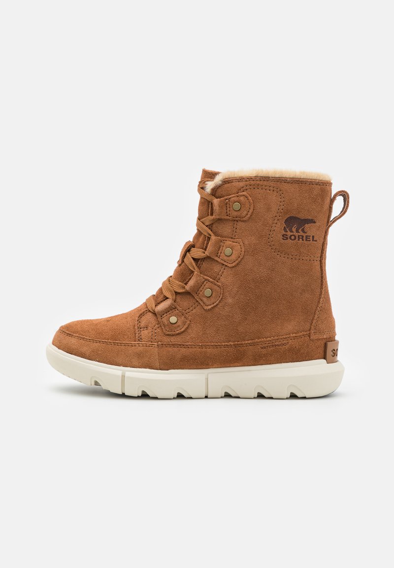 Botines de ante marrón con forro de pelo sintético, diseño de cordones, acentos en metal y suela de goma blanca. Presenta un logo en el lateral.