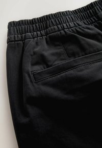 Fekete nadrág elasztikus derékkal és oldalsó zsebbel. Az anyag simának tűnik, matt felülettel, kiemelve a minimalista dizájnt.