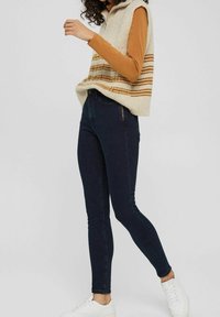 Kvinna som bär en beige ärmlös stickad väst med bruna randar över en senapsgul långärmad tröja, mörka skinny jeans och vita sneakers.