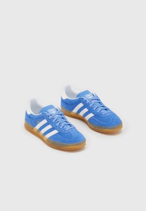 Par blå ruskind Adidas sneakers med hvide striber og gummisåler på en lysegrå baggrund.