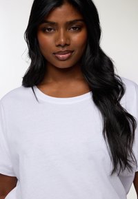 T-shirt bianco in cotone con collo rotondo, texture liscia e maniche corte, indossato da una persona con lunghi capelli mossi e scuri.