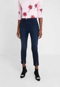 Jeans de mezclilla elásticos de color azul oscuro con un diseño ajustado. Presentan bolsillos frontales y aberturas en el dobladillo con acentos decorativos. Combinados con un suéter rosa adornado y botas negras hasta el tobillo.