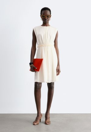 Frau trägt ärmelloses, cremefarbenes knielanges Kleid, braune Riemchenabsätze, braune Sonnenbrille, goldene Creolen, schwarzes Armband, hält eine rote Clutch.