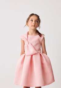 Abito rosa con un tessuto strutturato caratterizzato da motivi in rilievo. Il design include un dettaglio a fiocco e una gonna svasata, creando una silhouette giocosa.