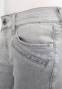Acercamiento de unos jeans de mezclilla gris claro que destacan el bolsillo delantero, el bolsillo para monedas, el lazo del cinturón y el cierre de botón de metal.