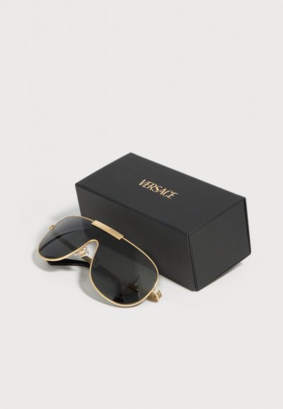 Μαύρα και χρυσά αεροπόρα γυαλιά ηλίου με σκουρόχρωμους φακούς δίπλα σε ένα μαύρο κουτί με το χρυσό λογότυπο "Versace" σε λευκό φόντο.