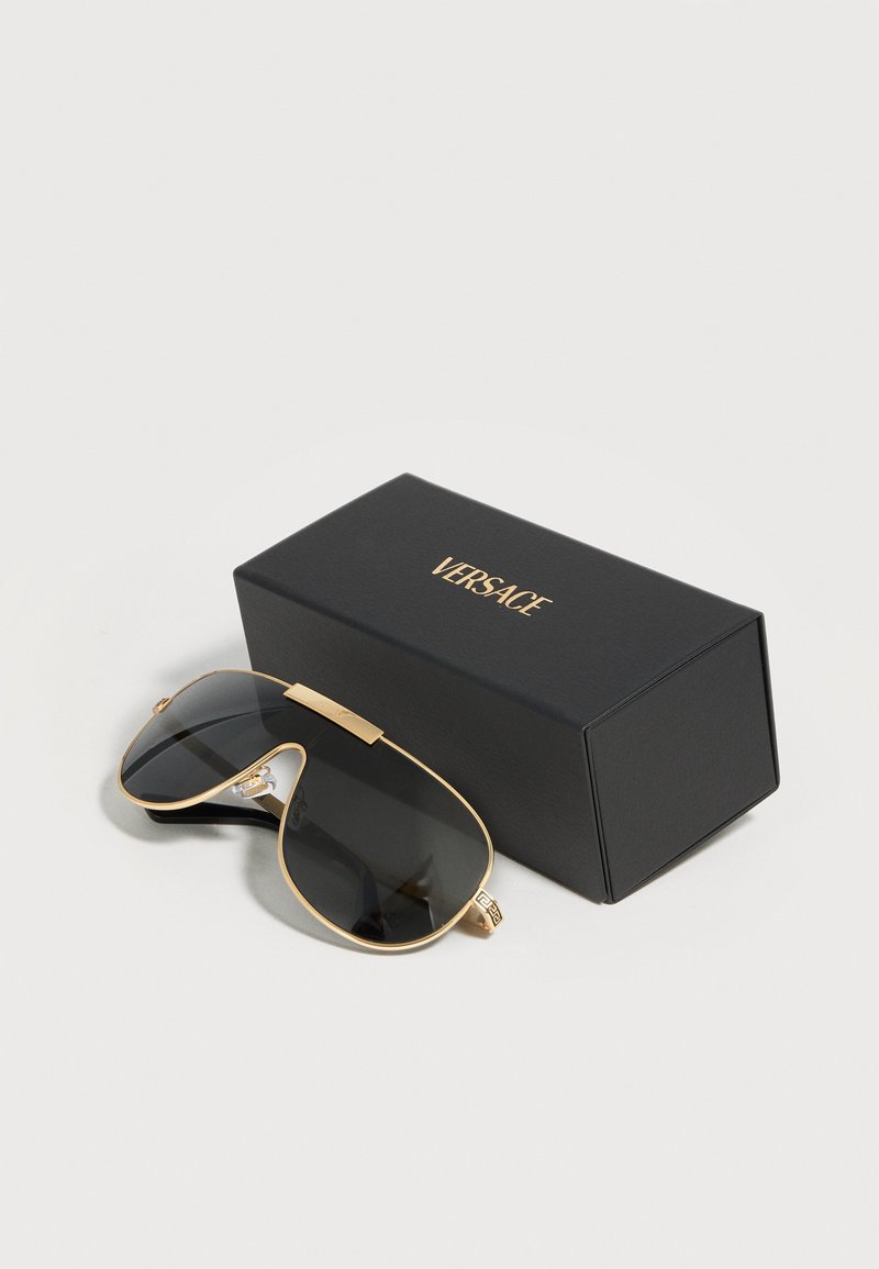 Black and gold aviator sunglasses with dark lenses next to a black box with gold "Versace" logo on a white background.