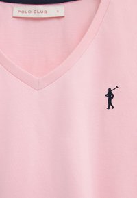 Camiseta de cuello en V color rosa claro con una pequeña figura negra bordada que lleva un mazo de polo en el lado superior izquierdo y una etiqueta "POLO CLUB" dentro del cuello.