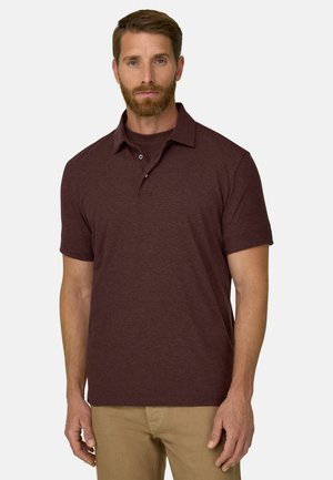 Mann mit Bart, der ein kurzärmeliges, kastanienbraunes Poloshirt und beige Hosen trägt, steht vor einem schlichten hellen Hintergrund.