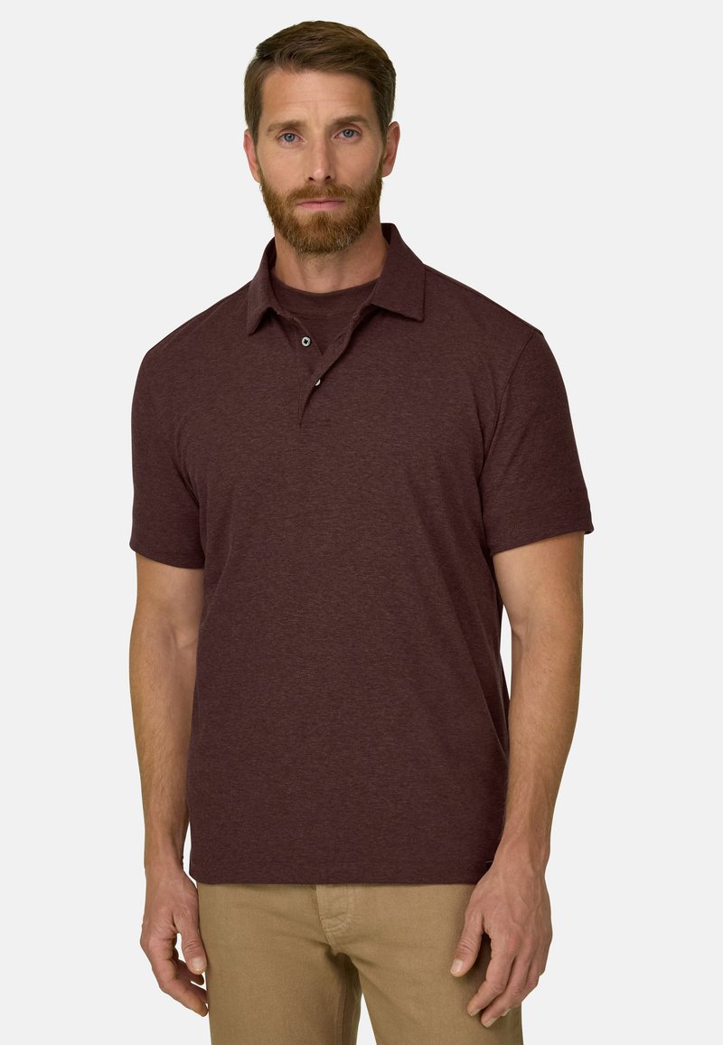 Homme barbu portant un polo bordeaux à manches courtes et un pantalon beige, debout devant un fond clair uni.