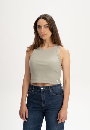 Femme aux longs cheveux bruns portant un crop top beige sans manches et un jean bleu foncé taille haute, debout devant un fond blanc uni.
