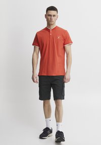 Rød polo med krage og hvite detaljer, sammen med svarte cargoshorts. Modellen har på seg svarte sneakers og hvite ankelstrømper.