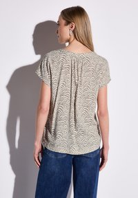 Kortærmet bluse i lys beige med et bølget mønster, med plisseret ryg og afslappet pasform, parret med mørkeblå jeans.