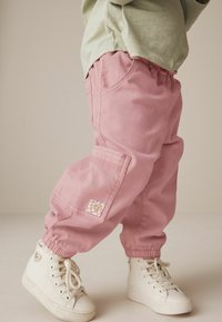 Pantaloni cargo din bumbac roz, cu manșete elastice, buzunare laterale și o aplicatie decorativă pe piciorul stâng. Împreună cu adidași albi cu toc înalt.