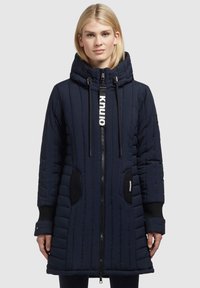 Navy quilted long coat with a high collar, zip closure, drawstring hood, and contrasting black pocket accents. Features textured fabric.