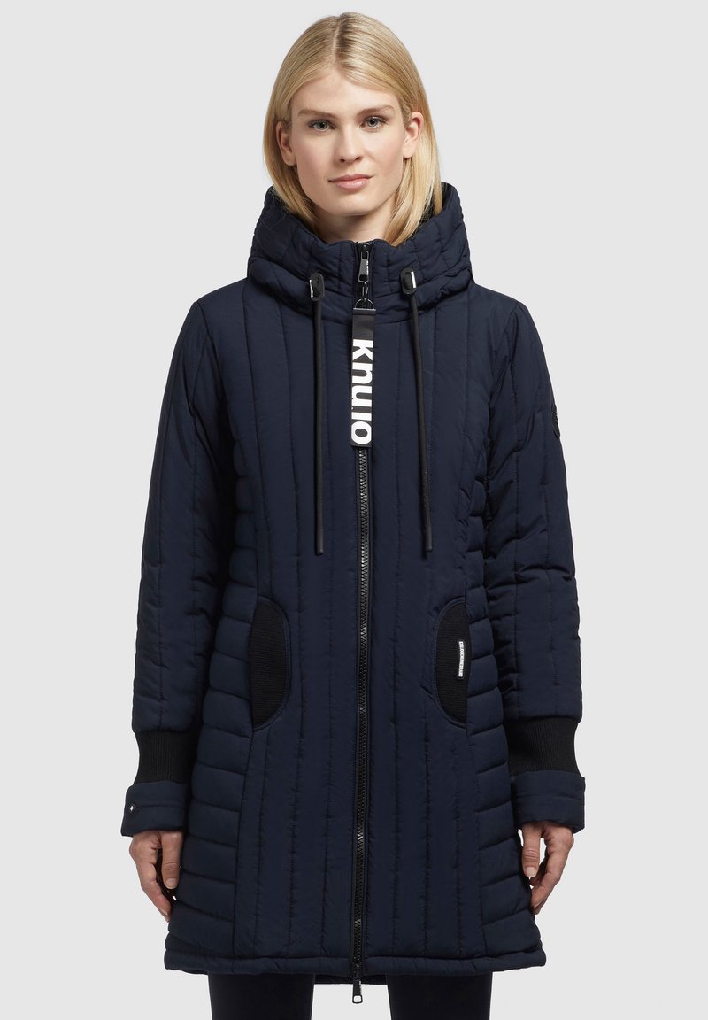 Navy quilted long coat with a high collar, zip closure, drawstring hood, and contrasting black pocket accents. Features textured fabric.