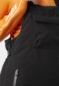 Overoles negros con un bolsillo lateral con cremallera y un bolsillo frontal con solapa. Fabricados con un material suave y resistente al agua, con tirantes ajustables.