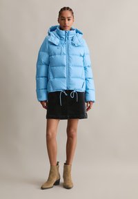 Helleblaue Steppjacke mit Kapuze, Reißverschluss, geripptem Saum und Bindebändern. Getragen über einem schwarzen Rock und hellbraunen Ankle Boots.