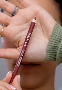 Czerwony ołówek do ust Isadora trzymany w dłoni, z ostro zakończonym końcem i nazwą marki wydrukowaną na białym tle. Teksturowany uchwyt blisko podstawy.