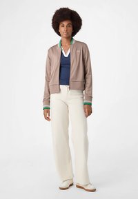 Brun zip-up jacka med gröna och vita randiga detaljer, lager-på-lager över en marinblå skjorta. Matchas med krämfärgade byxor och vita sneakers.