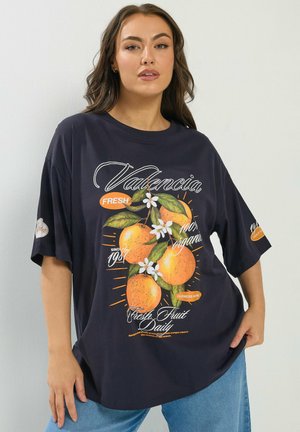 Kobieta w luźnej czarnej koszulce z grafiką pomarańczy z Walencji i napisem promującym świeże, ekologiczne owoce oraz targ rolniczy.
