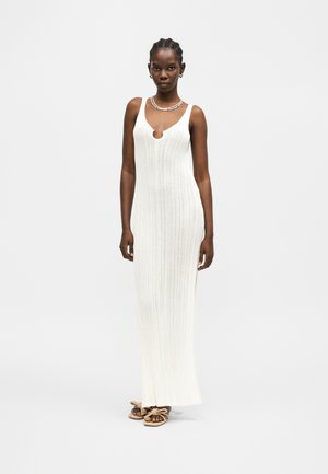 Naine, kes kannab valget varrukateta soonikutega maksikleit ümarate lõigeteeliste kaelusega, sandaale ja lühikesi punutisi, ühevärvilise taustaga.