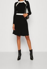 Vestido negro de manga larga con relieve, con acentos en blanco y botones dorados en los hombros, que cuenta con un dobladillo acampanado y se combina con botas negras de tobillo.