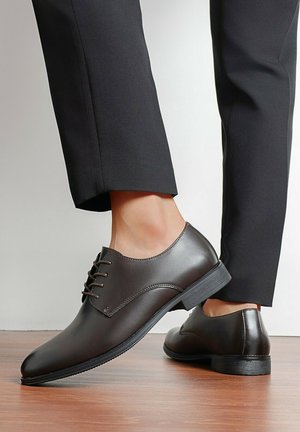 Chaussures Oxford en cuir marron à lacets portées avec un pantalon noir, présentées sur un sol en bois devant un fond uni.