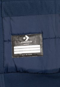 Pechera in tessuto blu navy con una superficie superiore testurizzata, caratterizzata da un'etichetta nera con testo bianco, logo e linee orizzontali.