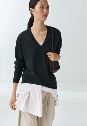 Donna con capelli scuri raccolti in una coda di cavallo, indossa un maglione nero con scollo a V, pantaloni beige e una camicia rosa chiaro legata in vita, tiene una piccola borsa marrone.