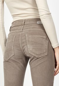 Brown corduroy pants with a slim fit, featuring two back pockets and subtle stitching details. It's paired with a light long-sleeve top.