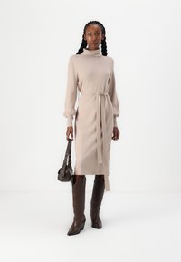 Abito lavorato a maglia beige con collo alto e vita cintrata, con maniche a sbuffo lunghe. Abbinato a stivali da cowboy marroni e una borsa piccola.