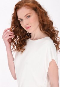 Witte gebreide top met een ontspannen pasvorm, korte mouwen en een zachte textuur. Model heeft golvend rood haar en een neutrale achtergrond.