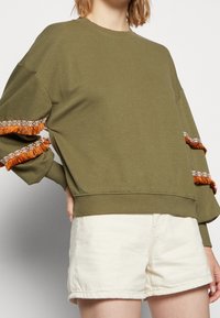 Sudadera verde oliva con acentos de flecos naranjas y estampados en las mangas y un dobladillo ajustado. Combinada con pantalones cortos blancos. Material de algodón.