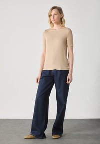 Haut beige à manches courtes avec un large décolleté associé à un pantalon en denim bleu foncé ample et des ballerines beige, sur une surface grise texturée.